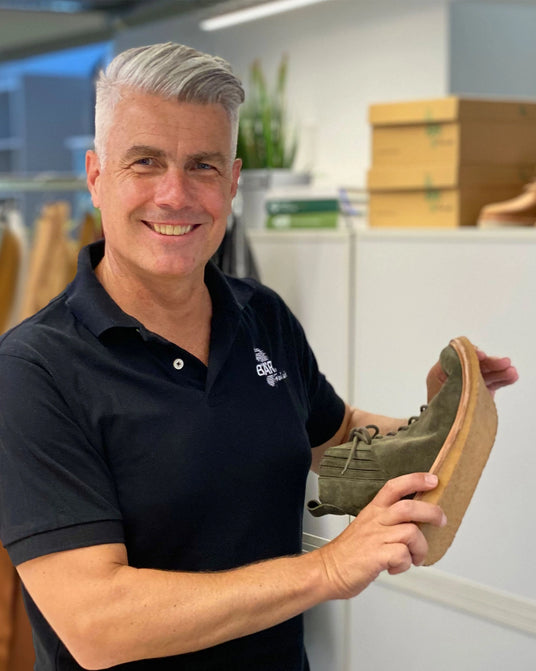Ein lächelnder Mann in schwarzem Polohemd hält einen grünen Schuh mit Korksohle in einem modernen Büro.
