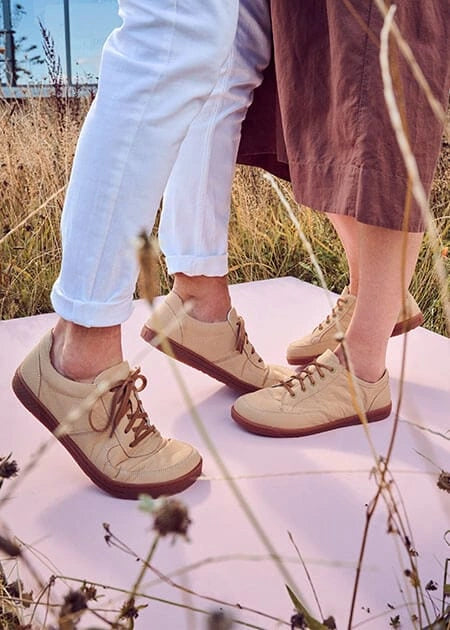 Zwei Frauen tragen helle, beige Sneaker und stehen auf einer rosafarbenen Plattform in einer sommerlichen Landschaft mit trockenem Gras.