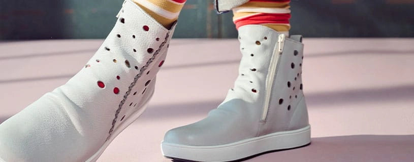 Nahaufnahme von weißen Damenstiefeletten mit schwarzen und roten Punkten. Eine Stiefelette steht auf einer hellen Oberfläche und hat einen seitlichen Reißverschluss, eine breite Zehenpartie und eine flache, helle Sohle. Im Hintergrund ist das Bein einer Person mit gestreiften Socken zu sehen, die eine zweite passende Stiefelette trägt.