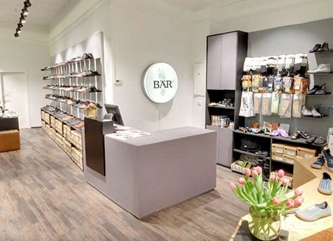 Innenansicht eines modernen Schuhladens mit Regalen voller Schuhe und Accessoires, einem grauen Tresen und einem Blumenstrauß auf einem Tisch