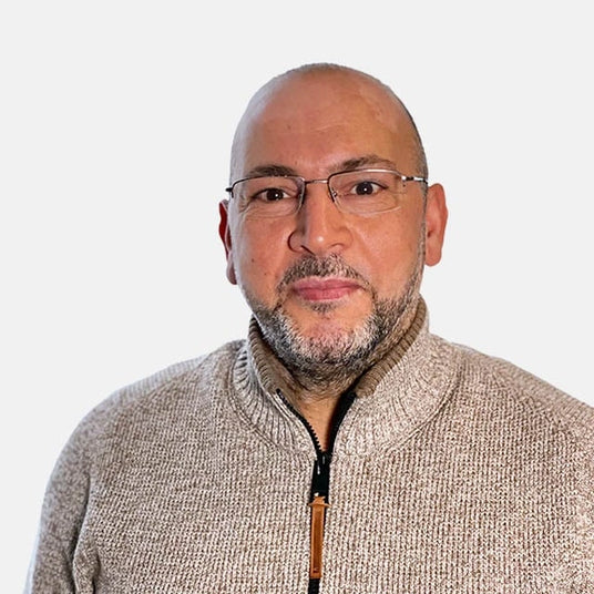 Ein Mann trägt eine Brille und einen beigen Pullover mit Reißverschluss, vor einem neutralen Hintergrund